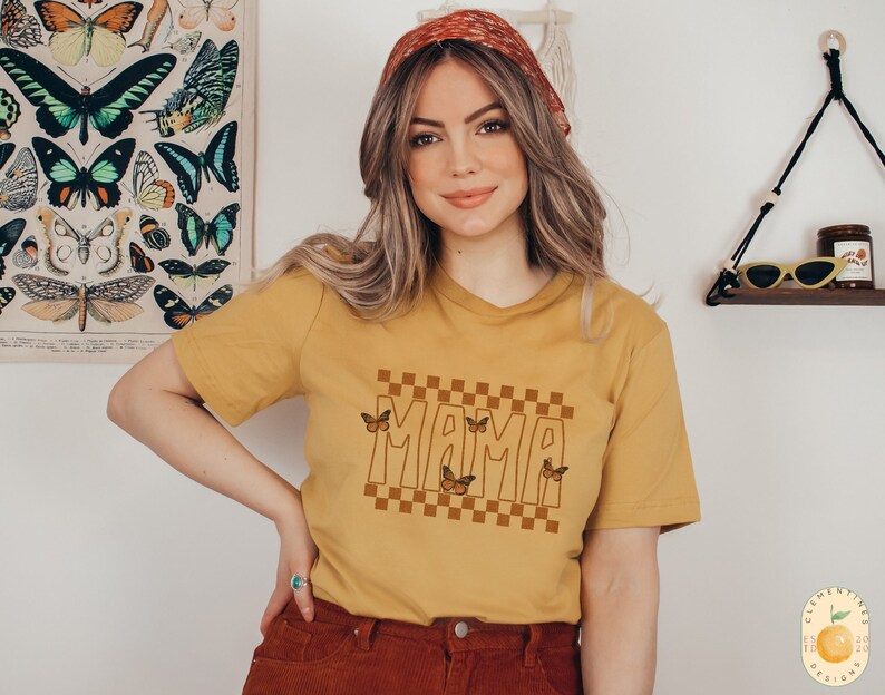 Puede incluir: Una camiseta amarilla mostaza con un patr&oacute;n a cuadros y la palabra "MAMA" en una fuente retro. Hay varios gr&aacute;ficos de mariposas marrones en la camiseta.