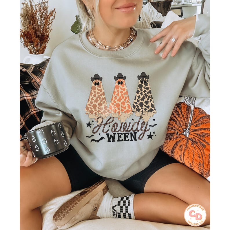 May include: A light gray sweatshirt featuring three ghost figures wearing cowboy hats with animal print patterns. The text "Howdy Ween" is below the ghosts, with stars and bats. A black mug with ghost designs is held in the hand.