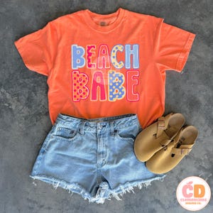 May include: An orange t-shirt with the words "BEACH BABE" in colorful block letters, paired with light-wash denim shorts and tan sandals. A casual summer outfit, perfect for a day at the beach.