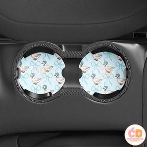 May include: Two black car cup holders with blue and white patterned inserts featuring a repeating design of white ducks wearing blue bonnets and blue bows.