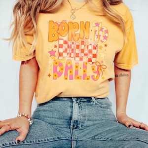 May include: Mustard yellow t-shirt with the text "BORN TO BUILD DALLYS" in orange and pink lettering. The design includes stars, a checkered pattern, and floral accents. The shirt is paired with blue jeans.