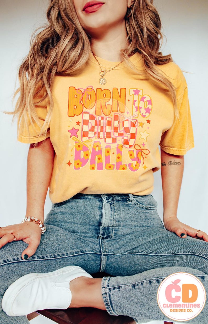 May include: Mustard yellow t-shirt with the text "BORN TO BUILD DALLYS" in orange and pink lettering. The design includes stars, a checkered pattern, and floral accents. The shirt is paired with blue jeans.