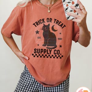 Op de afbeelding: Oranje-rood T-shirt met een zwarte afbeelding met een zwarte kat, een maan en de woorden "Trick or Treat Supply Co.". Het ontwerp bevat sterren en het jaar "1692". Het shirt maakt deel uit van een Halloween-collectie.