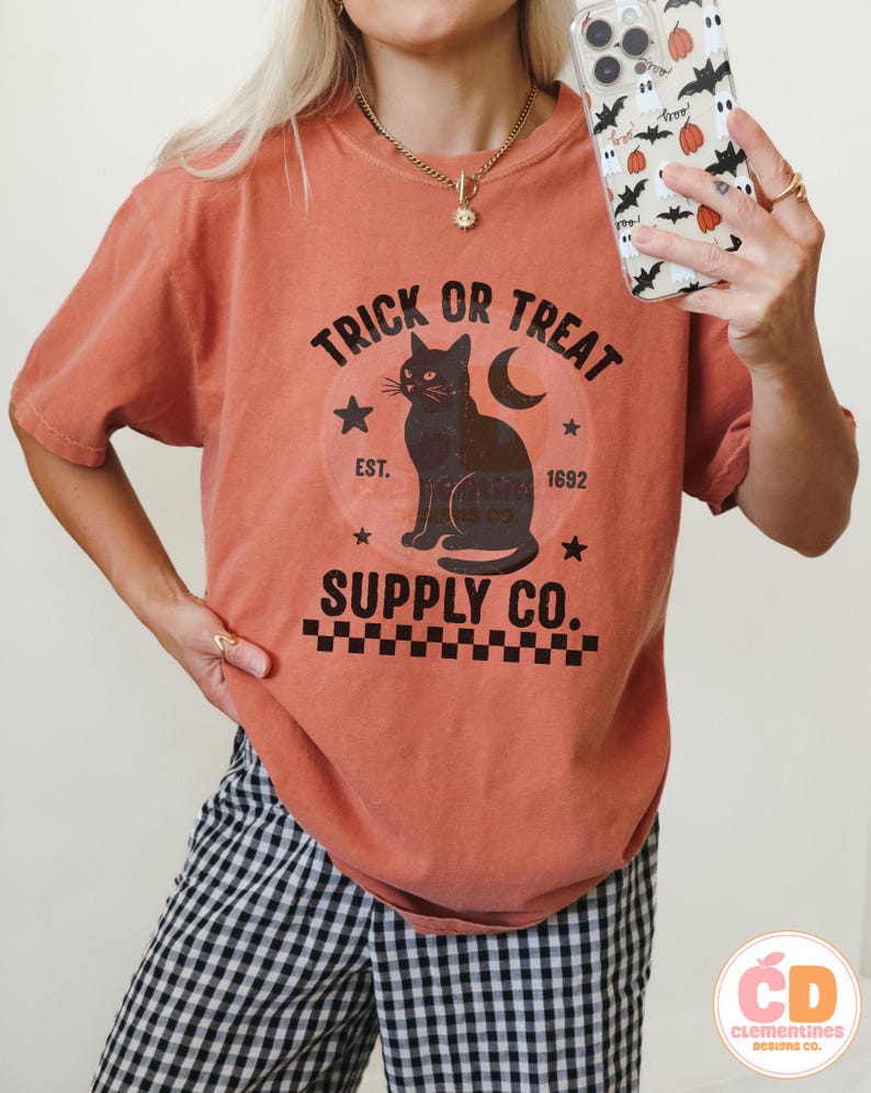 Op de afbeelding: Oranje-rood T-shirt met een zwarte afbeelding met een zwarte kat, een maan en de woorden "Trick or Treat Supply Co.". Het ontwerp bevat sterren en het jaar "1692". Het shirt maakt deel uit van een Halloween-collectie.