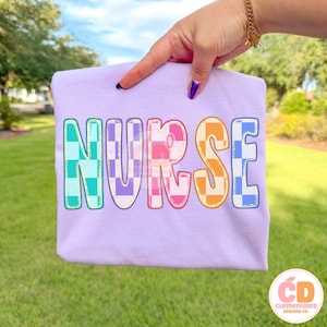 Pu&ograve; includere: Una maglietta lilla con la scritta "NURSE" in lettere colorate a scacchi. Le lettere sono in tonalit&agrave; di blu, viola, arancione e rosa. La maglietta &egrave; presentata su uno sfondo erboso verde.