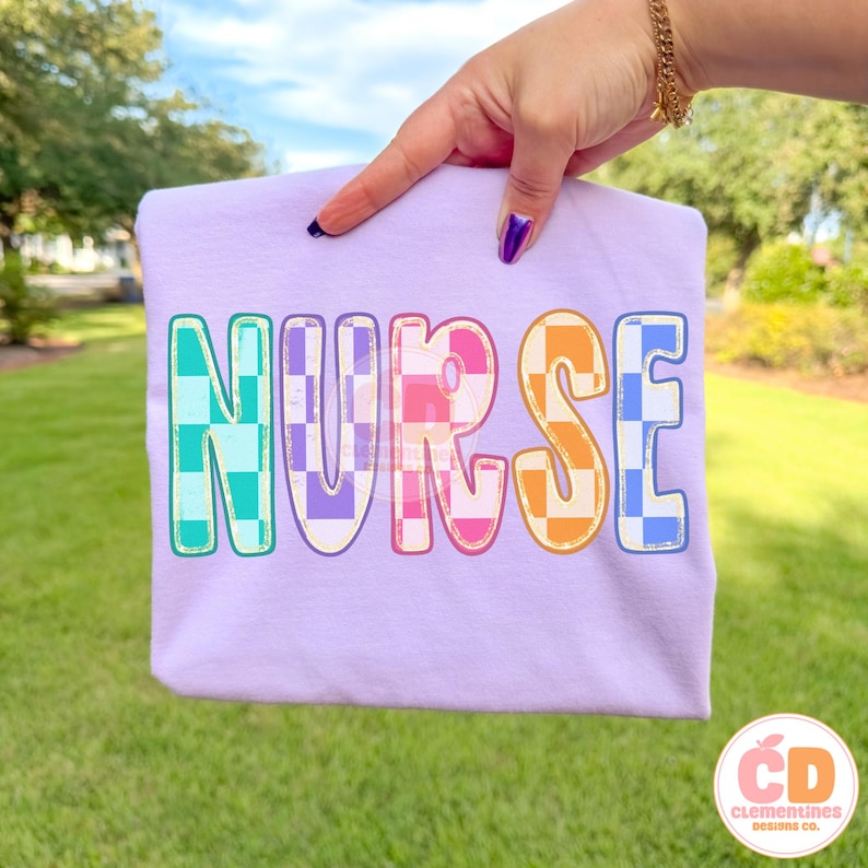 Pu&ograve; includere: Una maglietta lilla con la scritta "NURSE" in lettere colorate a scacchi. Le lettere sono in tonalit&agrave; di blu, viola, arancione e rosa. La maglietta &egrave; presentata su uno sfondo erboso verde.