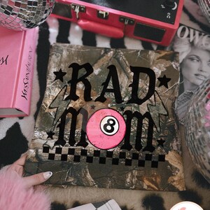 May include: A camouflage-patterned square fabric with the words "RAD MOM" in black script, a pink eight-ball graphic, and a black and white checkered border. The fabric is surrounded by a pink book, a record player, and disco balls.