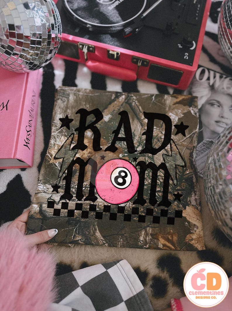 May include: A camouflage-patterned square fabric with the words "RAD MOM" in black script, a pink eight-ball graphic, and a black and white checkered border. The fabric is surrounded by a pink book, a record player, and disco balls.
