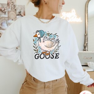 May include: White sweatshirt with a graphic of a goose wearing a blue bonnet and surrounded by flowers. The text "Mama Goose" is printed below the goose.