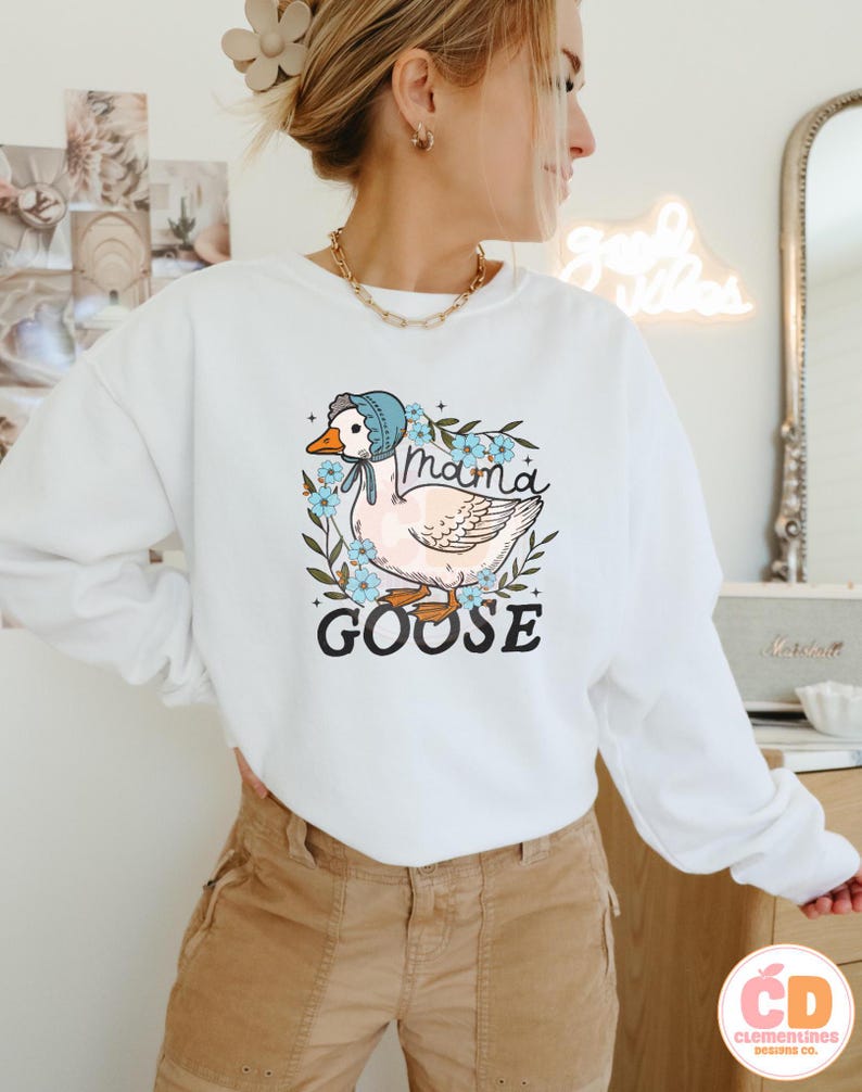May include: White sweatshirt with a graphic of a goose wearing a blue bonnet and surrounded by flowers. The text "Mama Goose" is printed below the goose.