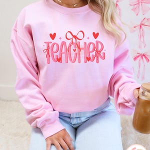 May include: Pink sweatshirt with the word "TEACHER" in red and white lettering with hearts and bows.