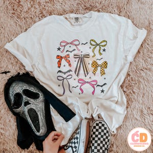 May include: Cream-colored t-shirt featuring a colorful bow graphic with bats and stars. A black and silver Scream mask and checkered Vans sneakers are also visible. The shirt has a relaxed fit, perfect for Halloween.