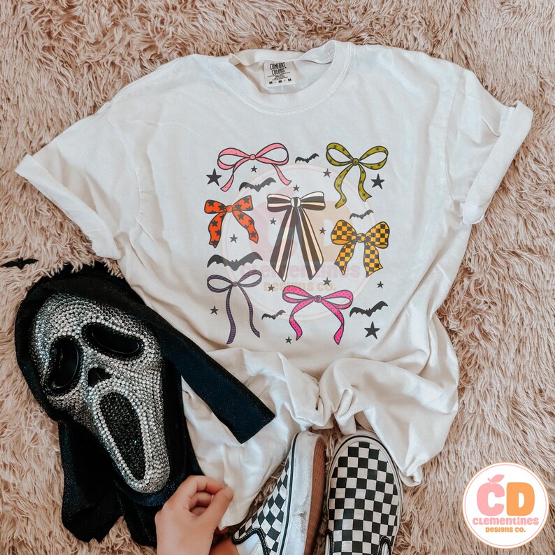 May include: Cream-colored t-shirt featuring a colorful bow graphic with bats and stars. A black and silver Scream mask and checkered Vans sneakers are also visible. The shirt has a relaxed fit, perfect for Halloween.