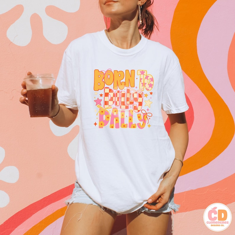 May include: White t-shirt with the text "BORN TO DOLLY DALLY" in orange, pink, and yellow. The design includes stars and a checkered pattern. The shirt is paired with denim shorts.
