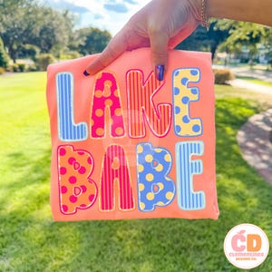 May include: A coral-colored t-shirt with the words "LAKE BABE" in colorful, patterned letters. The letters are outlined in a contrasting color, with polka dots and stripes. The shirt is held up by a hand.