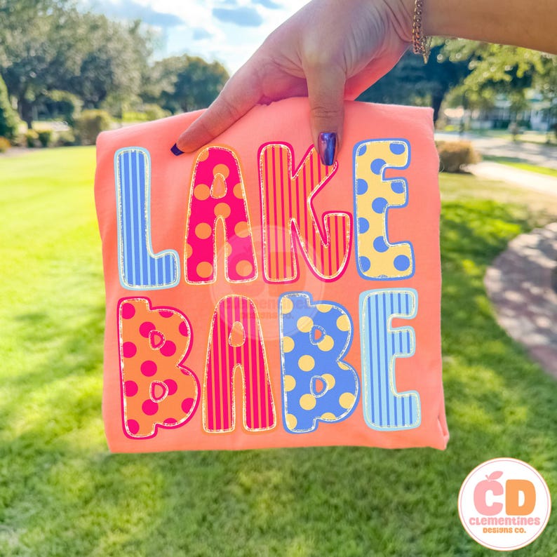 May include: A coral-colored t-shirt with the words "LAKE BABE" in colorful, patterned letters. The letters are outlined in a contrasting color, with polka dots and stripes. The shirt is held up by a hand.