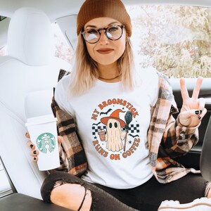 May include: White t-shirt with a Halloween ghost graphic wearing a witch hat, holding a broom and coffee. The text "MY BROOMSTICK RUNS ON COFFEE" is printed around the ghost. The person is wearing a brown beanie, glasses, and a plaid shirt.