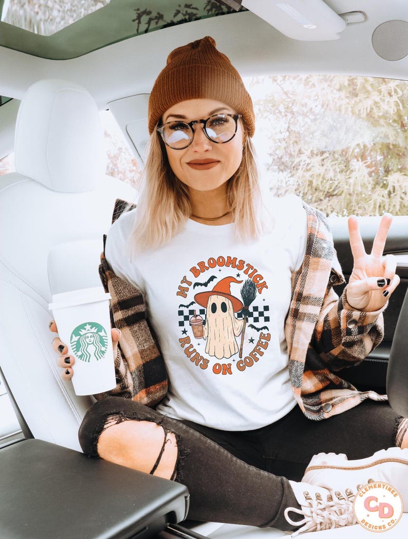May include: White t-shirt with a Halloween ghost graphic wearing a witch hat, holding a broom and coffee. The text "MY BROOMSTICK RUNS ON COFFEE" is printed around the ghost. The person is wearing a brown beanie, glasses, and a plaid shirt.