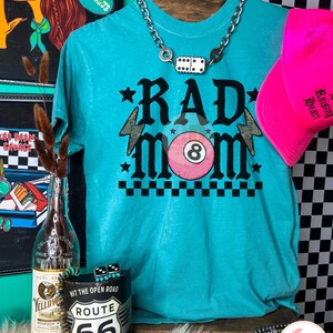 May include: A turquoise t-shirt with the words "RAD MOM" in black, featuring a pink 8-ball and lightning bolts. A pink baseball cap and a silver chain necklace with dice are also visible. The shirt is displayed with other accessories.
