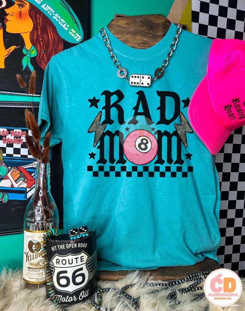 May include: A turquoise t-shirt with the words "RAD MOM" in black, featuring a pink 8-ball and lightning bolts. A pink baseball cap and a silver chain necklace with dice are also visible. The shirt is displayed with other accessories.