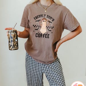 May include: A brown t-shirt with the text "FRESHLY BOO'D COFFEE" and a ghost graphic holding a coffee cup. The shirt is paired with black and white checkered pants. A leopard print tumbler with a straw is held in the hand.