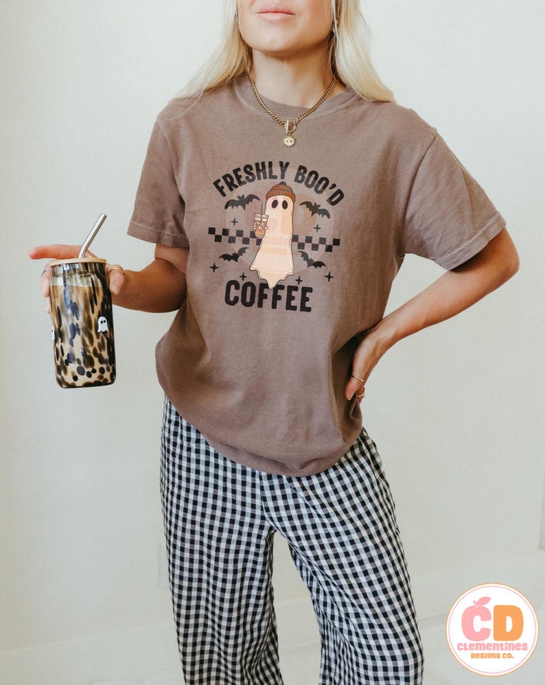 May include: A brown t-shirt with the text "FRESHLY BOO'D COFFEE" and a ghost graphic holding a coffee cup. The shirt is paired with black and white checkered pants. A leopard print tumbler with a straw is held in the hand.