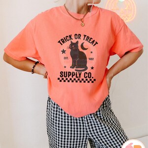 Op de afbeelding: Koraalrood T-shirt met een zwarte afbeelding van een kat, maan en sterren, met de tekst "TRICK OR TREAT SUPPLY CO." Het ontwerp heeft een vintage uitstraling, ideaal voor Halloween. Het model draagt een zwart-wit geruite broek.