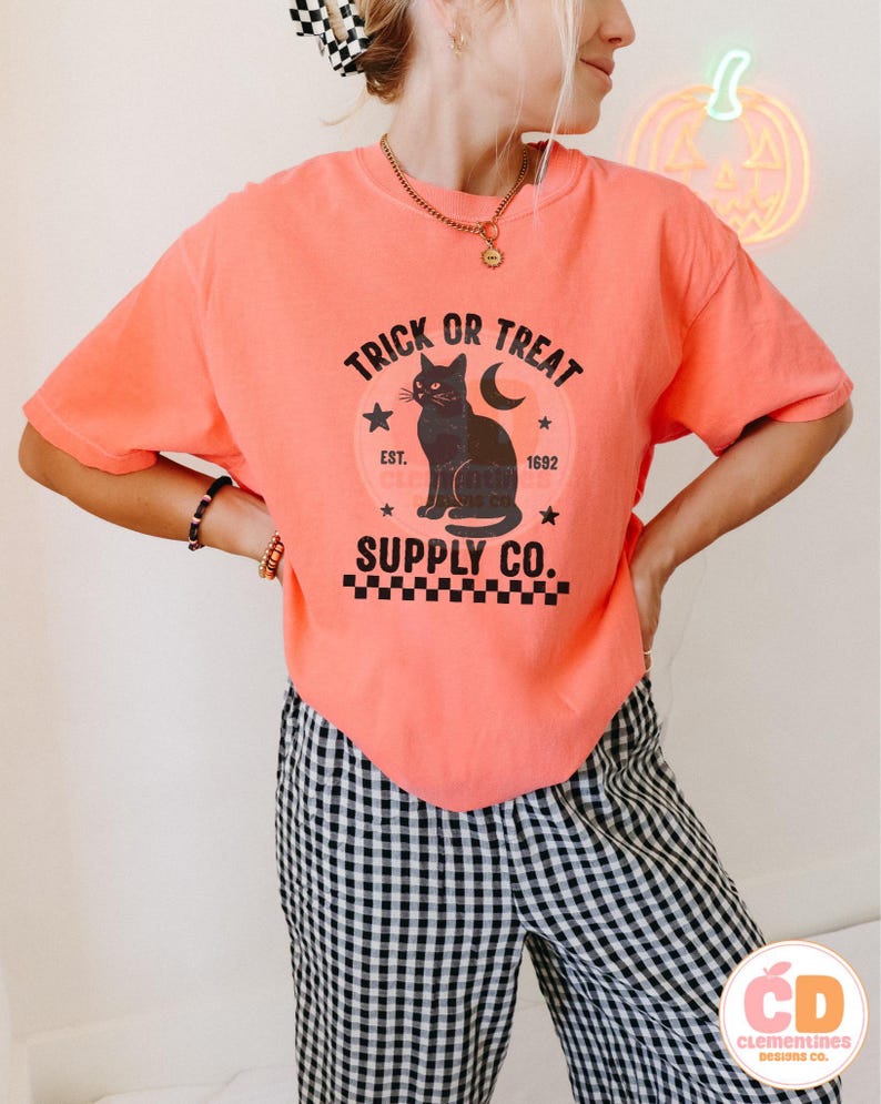 Op de afbeelding: Koraalrood T-shirt met een zwarte afbeelding van een kat, maan en sterren, met de tekst "TRICK OR TREAT SUPPLY CO." Het ontwerp heeft een vintage uitstraling, ideaal voor Halloween. Het model draagt een zwart-wit geruite broek.