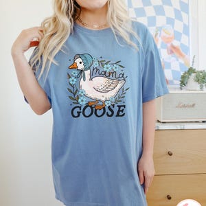 May include: A light blue t-shirt with a graphic of a white goose wearing a bonnet and surrounded by blue flowers. The text "mama goose" is printed below the goose.