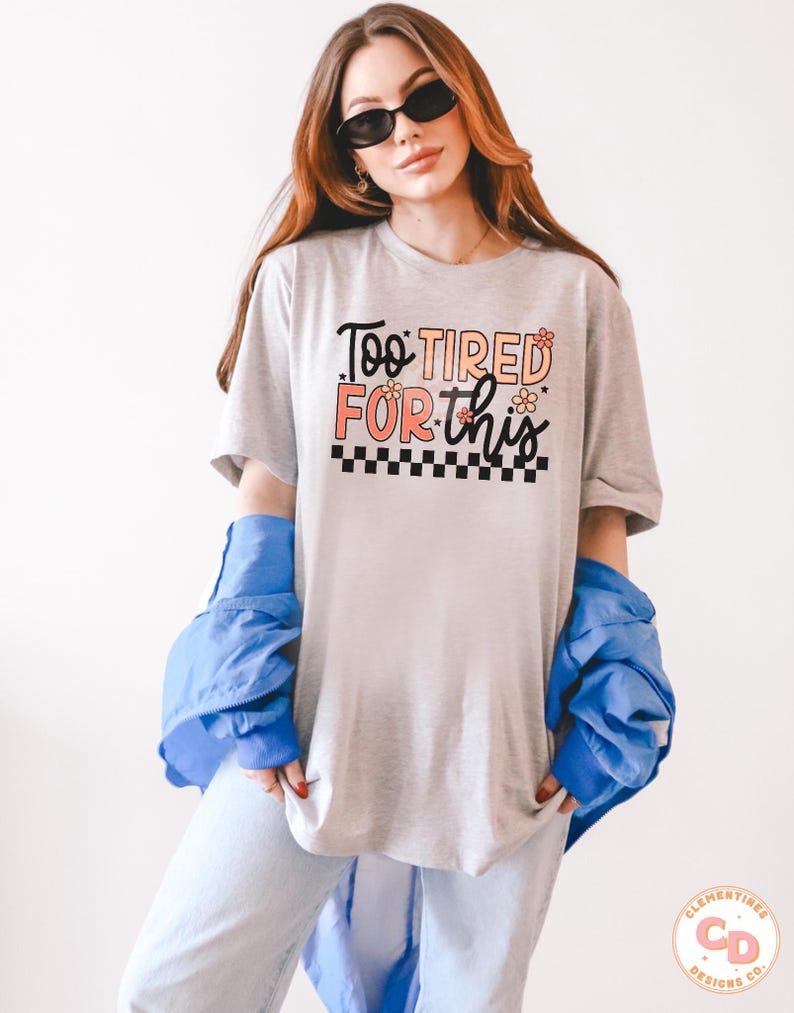May include: A gray t-shirt with the text "Too Tired For This" in black and orange lettering with small flower accents. The text is surrounded by a black and white checkered pattern.
