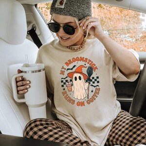 May include: Beige t-shirt with a Halloween ghost graphic that says "My Broomstick Runs on Coffee." The ghost wears a witch hat and holds a broom and coffee. The person is wearing a grey beanie and sunglasses.