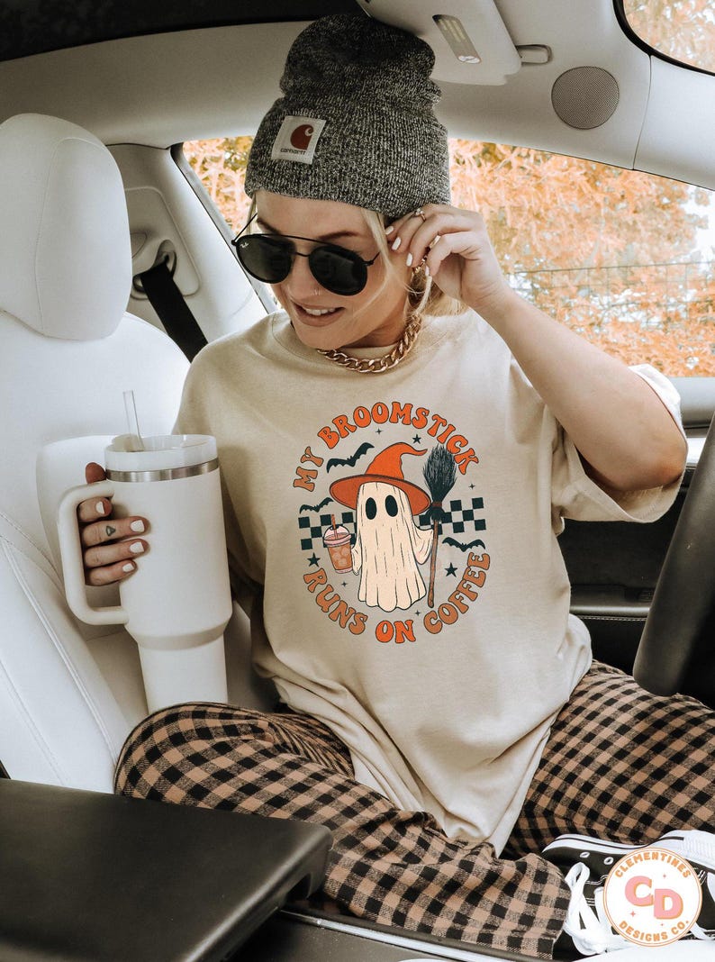 May include: Beige t-shirt with a Halloween ghost graphic that says "My Broomstick Runs on Coffee." The ghost wears a witch hat and holds a broom and coffee. The person is wearing a grey beanie and sunglasses.