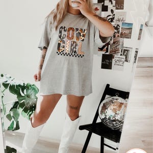 Puede incluir: Una camiseta gris con las palabras "COW GIRL" en una mezcla de fuentes y patrones. La camiseta se combina con botas de vaquero blancas y una gorra de b&eacute;isbol negra. Una bola de discoteca est&aacute; sobre una silla negra.