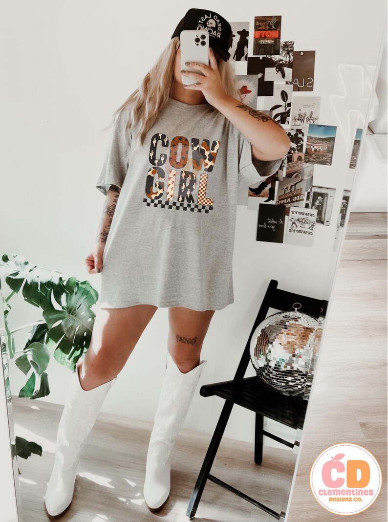 Puede incluir: Una camiseta gris con las palabras "COW GIRL" en una mezcla de fuentes y patrones. La camiseta se combina con botas de vaquero blancas y una gorra de b&eacute;isbol negra. Una bola de discoteca est&aacute; sobre una silla negra.