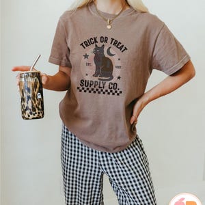 Op de afbeelding: Een bruin t-shirt met de tekst "TRICK OR TREAT SUPPLY CO." en een zwarte kattenafbeelding. Het shirt wordt gecombineerd met een zwart-wit geruite broek. Een beker met een rietje en een luipaardprint wordt in de hand gehouden.