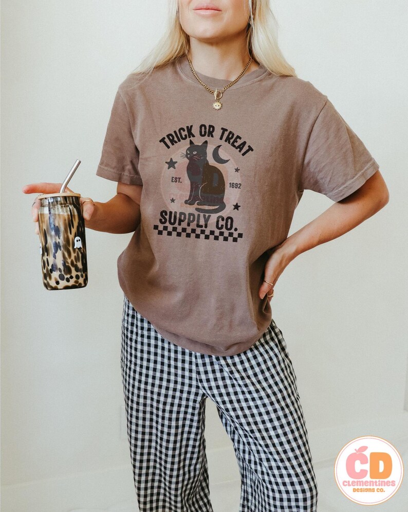 Op de afbeelding: Een bruin t-shirt met de tekst "TRICK OR TREAT SUPPLY CO." en een zwarte kattenafbeelding. Het shirt wordt gecombineerd met een zwart-wit geruite broek. Een beker met een rietje en een luipaardprint wordt in de hand gehouden.