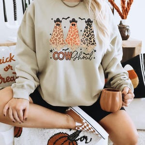 May include: A light beige sweatshirt with a graphic of three ghosts wearing cowboy hats and a "COW GHOUL" text design. The ghosts are wearing a brown, orange, and black cow print pattern.