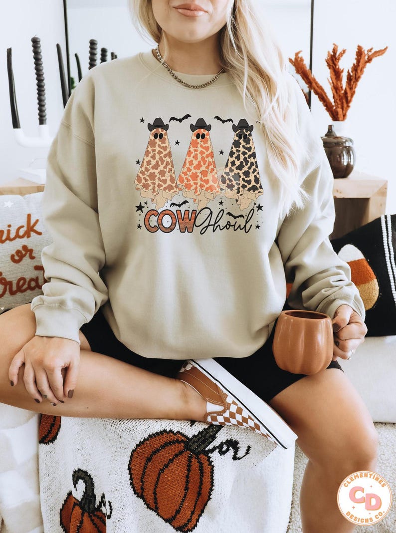 May include: A light beige sweatshirt with a graphic of three ghosts wearing cowboy hats and a "COW GHOUL" text design. The ghosts are wearing a brown, orange, and black cow print pattern.