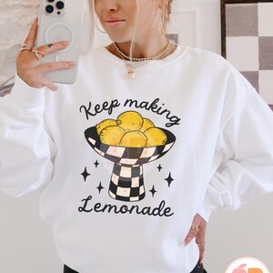 May include: White sweatshirt with a graphic of lemons in a black and white checkered bowl, with the text "Keep making Lemonade". The design includes black star accents. The sweatshirt is a casual, long-sleeved style.