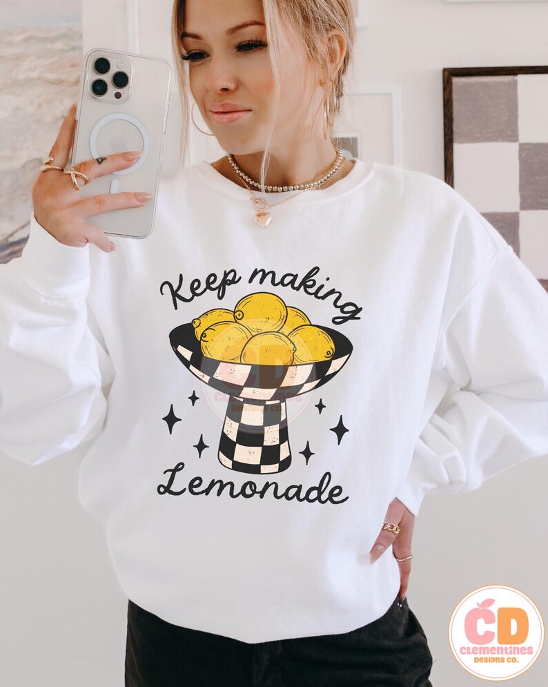 May include: White sweatshirt with a graphic of lemons in a black and white checkered bowl, with the text "Keep making Lemonade". The design includes black star accents. The sweatshirt is a casual, long-sleeved style.