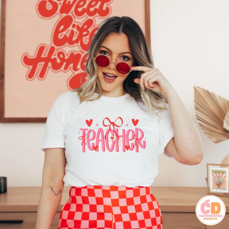May include: White t-shirt with pink text that says "TEACHER" with a pink bow and hearts. The model is wearing red and white checkered pants and red sunglasses.
