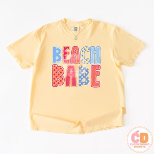 May include: A light yellow t-shirt with the words "BEACH BABE" in colorful, patterned letters. The shirt has short sleeves and a crew neck. A silver necklace is visible at the neckline. The shirt is from Clementines Designs Co.