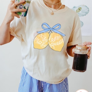 以下が含まれることがあります： 青いリボンで結ばれた2つのレモンのグラフィックが描かれた薄いベージュのTシャツ。レモンには「easy peasy」と「lemon squeezy」の文字が書かれています。