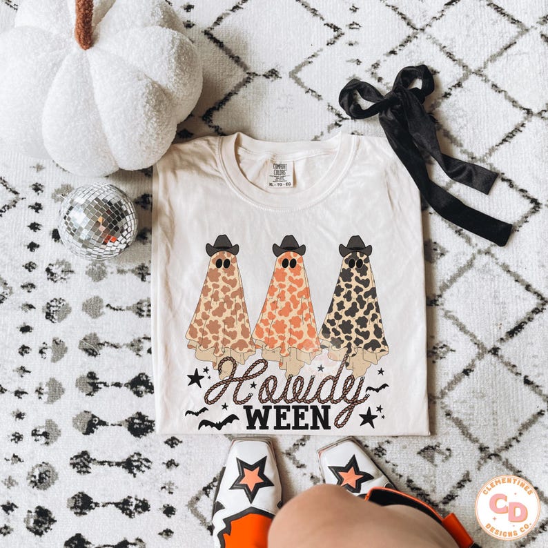 May include: Cream-colored t-shirt featuring three ghost figures wearing cowboy hats, with cow-print patterns in brown, orange, and black. The text "Howdy Ween" is written in a script font below the ghosts, with stars and bats. A white pumpkin and disco ball are also in the image.