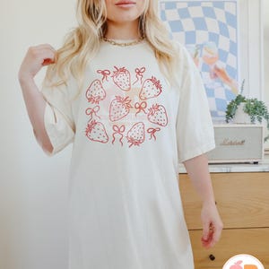 K&ouml;nnte beinhalten: Ein wei&szlig;es Oversize-T-Shirt mit einem Motiv aus Erdbeeren mit roten Schleifen in einem Kreis. Das Shirt hat eine lockere Passform und kurze &Auml;rmel.