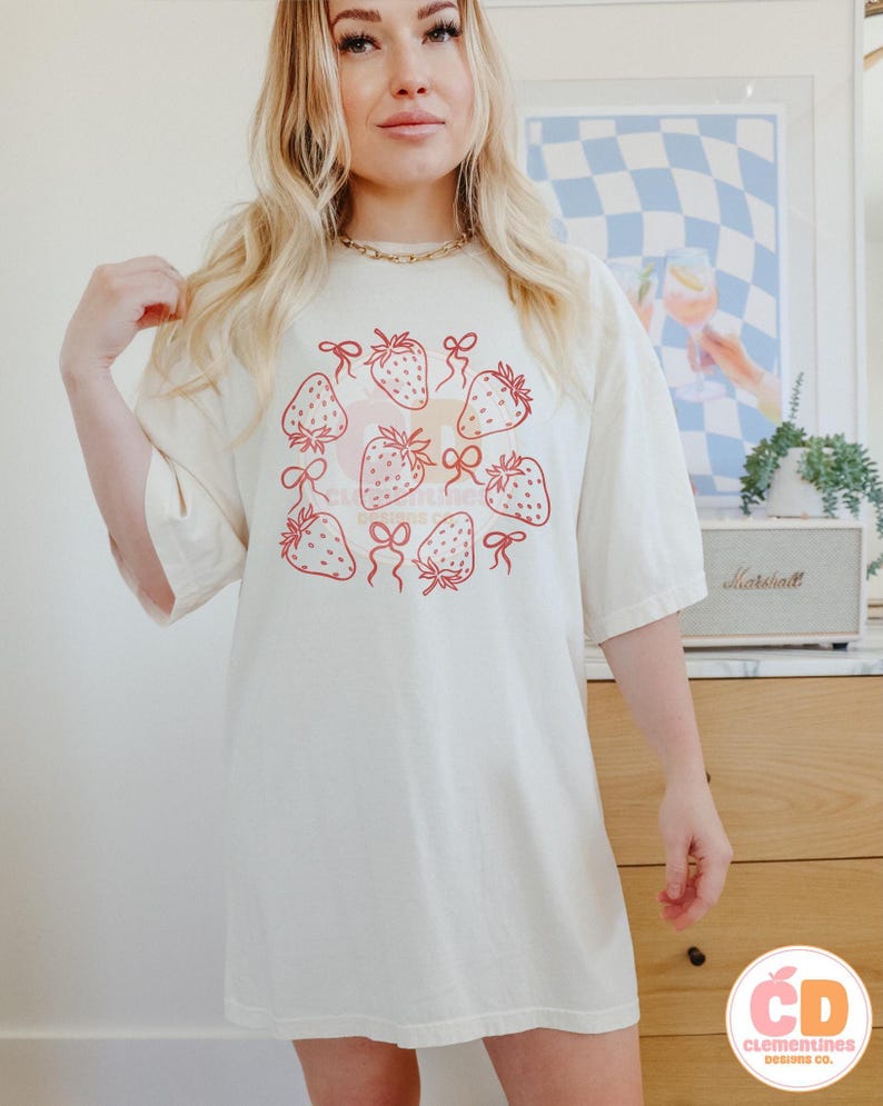K&ouml;nnte beinhalten: Ein wei&szlig;es Oversize-T-Shirt mit einem Motiv aus Erdbeeren mit roten Schleifen in einem Kreis. Das Shirt hat eine lockere Passform und kurze &Auml;rmel.