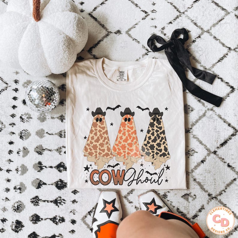 May include: A white t-shirt with a graphic of three cow ghosts wearing cowboy hats. The ghosts are wearing a brown, orange, and black cow print. The text "COW Ghoul" is printed below the ghosts.