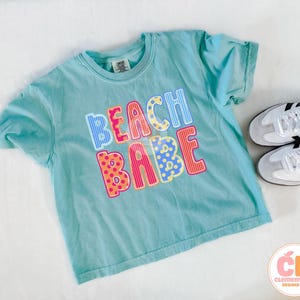 May include: A turquoise t-shirt with the words "BEACH BABE" in colorful, patterned letters. The shirt is paired with white sneakers with black stripes. The shirt is from Clementines Designs Co.