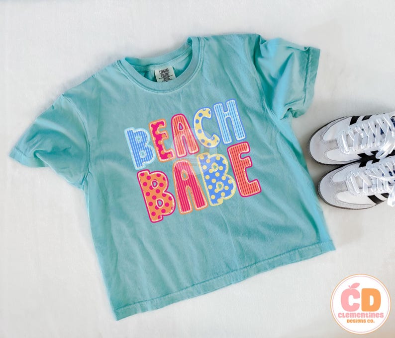 May include: A turquoise t-shirt with the words "BEACH BABE" in colorful, patterned letters. The shirt is paired with white sneakers with black stripes. The shirt is from Clementines Designs Co.