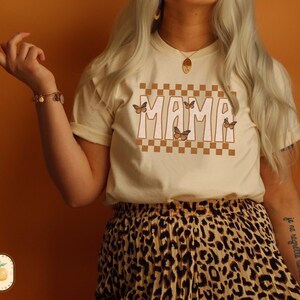 Puede incluir: Una camiseta de color crema con un patr&oacute;n a cuadros y la palabra "MAMA" en fuente marr&oacute;n. La camiseta tiene gr&aacute;ficos de mariposas. La modelo lleva una falda de estampado de leopardo.